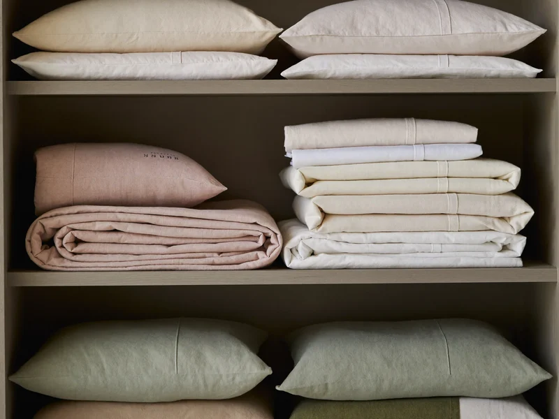 Draps et taies pliés en tons argile, crème et sauge sur une étagère en bois